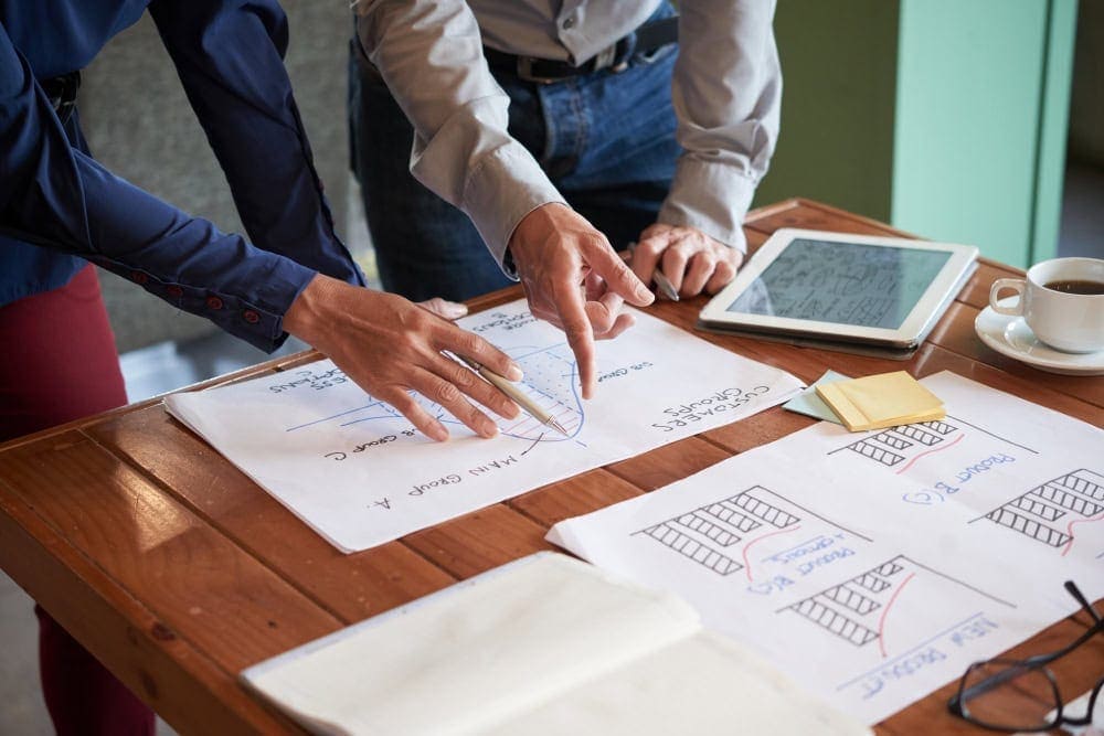 Business professionals collaborating on project plans at a wooden table with sketches, tablet, and coffee, emphasizing teamwork and strategic planning.