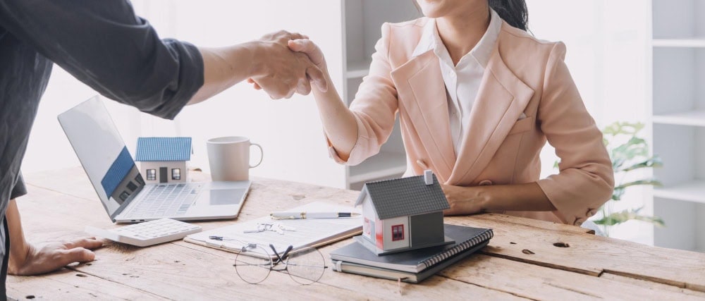 Professional real estate agent shaking hands with client in office, discussing property investment or sale, with house models and laptop on desk.