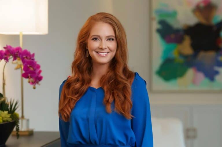 Portrait of a smiling woman with long red hair wearing a blue blouse in a modern interior setting in Frisco, TX.