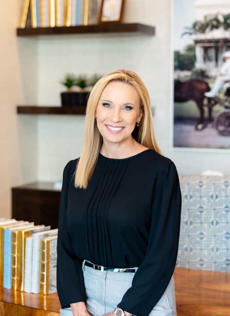Smiling professional woman with blonde hair in a modern office, wearing a black blouse and light gray pants, with books and decor in the background.